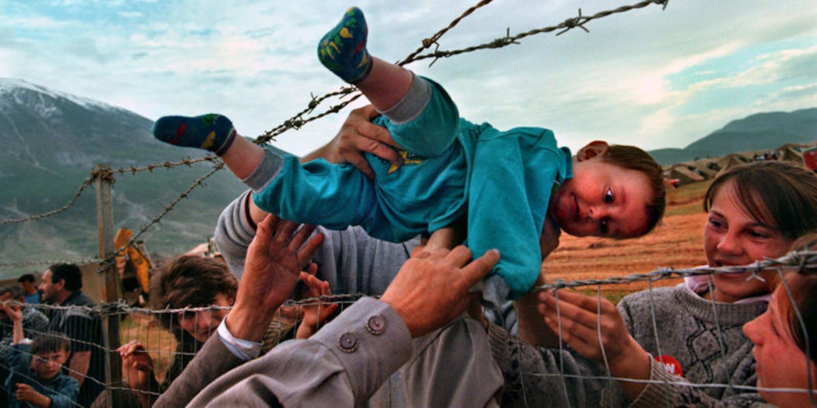 Agim Shala, de 2 anos, é passado por uma cerca de arame farpado para os braços de seus avós no campo de refugiados de Kukes, Albânia, em 3 de maio de 1999. A foto, tirada por Carol Guzy, do The Washington Post, ganhou o Prêmio Pulitzer e tornou-se um símbolo da crise de refugiados do Kosovo. Fonte: Carol Guzy / The Washington Post.