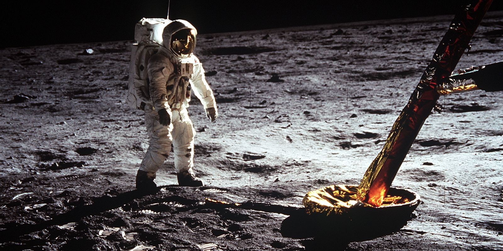 Buzz Aldrin é fotografado na superfície lunar, com a bandeira dos Estados Unidos ao fundo e o Módulo Lunar Eagle parcialmente visível. A imagem reflete a vastidão do ambiente lunar e a presença humana em um novo mundo. Fonte: NASA / Neil Armstrong.