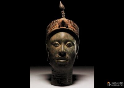Cabeça de bronze de Ifé, criada pelo povo Yoruba na Nigéria entre os séculos XII e XIII. A escultura, feita com técnica de cera perdida, mede aproximadamente 35 centímetros e apresenta naturalismo excepcional. O rosto é coberto por linhas verticais incisas, enquanto a coroa elaborada possui múltiplas camadas de contas tubulares e borlas. Restos de tinta vermelha e preta ainda são visíveis na superfície, indicando que originalmente era policromada. Fonte: © Anthony Denselow / BBC