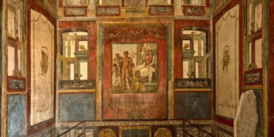 Vista geral do interior da Casa dos Vettii mostrando a arquitetura romana preservada. Os afrescos cobrem todas as paredes com painéis mitológicos emoldurados. A erupção do Vesúvio em 79 d.C. selou a casa sob cinzas, preservando-a por quase 2 mil anos.