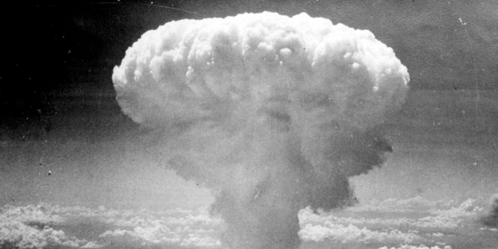 O cogumelo atômico subindo sobre Hiroshima em 6 de agosto de 1945. Esta imagem icônica, capturada a uma distância segura, mostra a imensa nuvem de fumaça, poeira e detritos que se formou após a detonação da bomba "Little Boy". A nuvem atingiu uma altitude de mais de 12 quilômetros, tornando-se um símbolo visual da Era Atômica. Fonte: UN Photo.