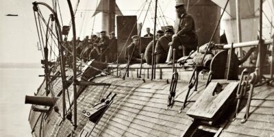 USS Galena após a Batalha de Drewry's Bluff em 15 de maio de 1862. A fotografia mostra o convés perfurado com três grandes buracos visíveis no casco de madeira. Tripulantes caminham entre os destroços enquanto canhões Dahlgren permanecem em posição. O navio foi atingido 44 vezes durante o bombardeio de Fort Darling, expondo a inadequação de sua blindagem de apenas 79 milímetros. Esta imagem histórica captura o momento em que a Marinha dos EUA aprendeu lições dolorosas sobre proteção naval. Fonte: James F. Gibson / Library of Congress / U.S. Naval Historical Center