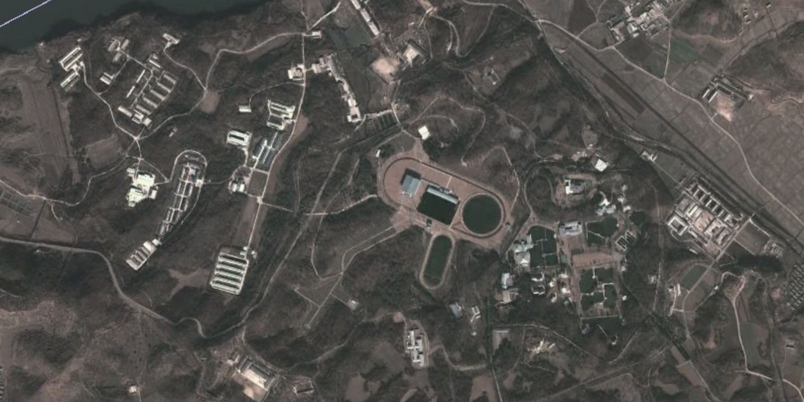 Vista aérea de satélite mostra a extensão do complexo Kang Dong com seus lagos artificiais e jardins elaborados. Construído na década de 1980 e expandido nos anos 90 sob ordem de Kim Jong Il, o complexo custou estimados 140 milhões de dólares. Durante a Grande Fome da década de 1990, quando centenas de milhares de norte-coreanos morriam, Kang Dong continuava recebendo expansões e melhorias luxuosas.Fonte: AccessDPRK / Google Earth