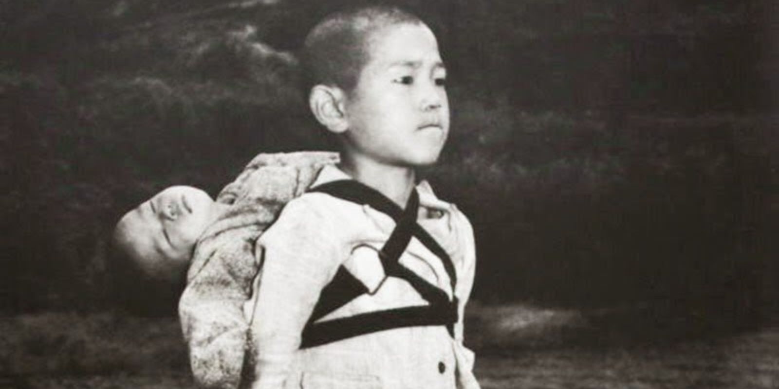 Um close-up do menino de Nagasaki, destacando sua expressão estoica e a determinação em seu olhar. Ele morde o lábio inferior com força, um sinal da imensa dor e responsabilidade que ele carrega em silêncio. A imagem foca na resiliência de uma criança forçada a amadurecer em um instante. Autor: Joe O'Donnell