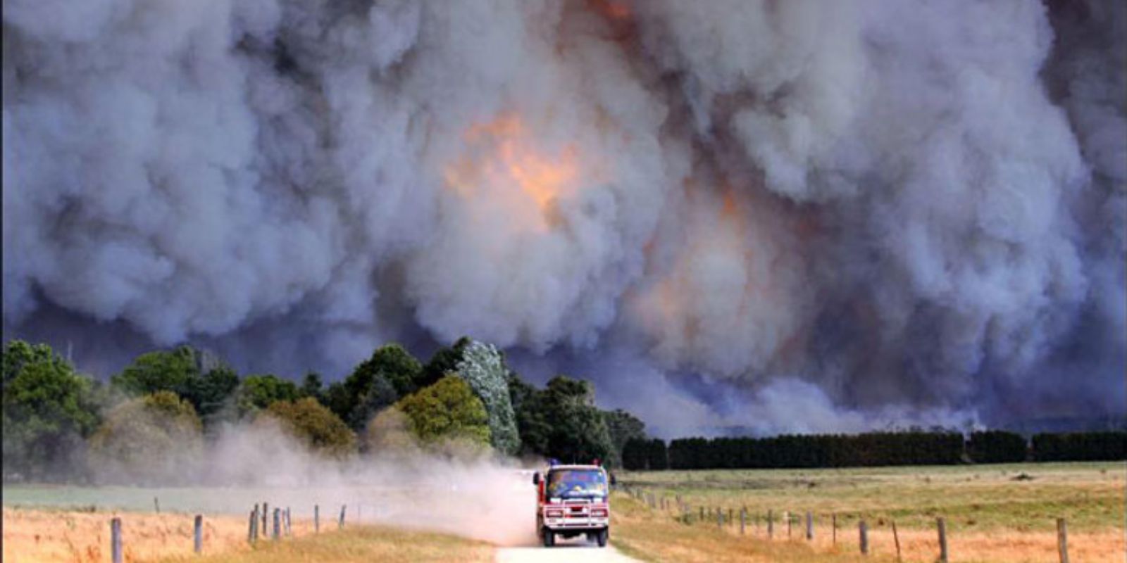 Nuvem massiva de fumaça e chamas durante os incêndios do Sábado Negro em Victoria, Austrália. Um veículo de emergência é visto fugindo da tempestade de fogo que se aproxima, ilustrando a velocidade e intensidade com que os incêndios se espalharam.