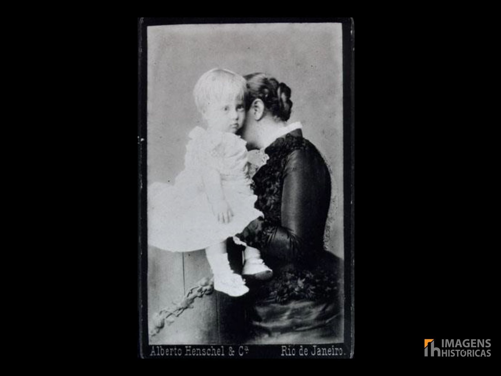 O retrato de 1882 mostra a Princesa Isabel sentada, segurando seu filho D. Antônio Gastão. Isabel veste um vestido elegante, típico da moda da época, e exibe uma expressão afetuosa e serena, enquanto o pequeno Antônio está nos braços da mãe, ainda um bebê. A composição é simples, mas eficaz, destacando o vínculo materno em um ambiente formal, típico dos retratos de estúdio da época.