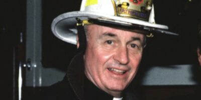 Padre Mychal Judge, capelão do Departamento de Bombeiros de Nova York (FDNY), fotografado com o capacete de bombeiro e sorriso característico. Judge dedicou sua vida a servir bombeiros, sem-teto, pacientes de AIDS e pessoas marginalizadas. Conhecido por trabalhar 16 horas por dia, ele era considerado um santo vivo antes de sua morte em 11 de setembro de 2001. Fonte: National Catholic Reporter