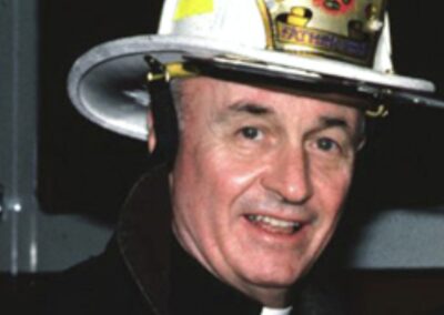 Padre Mychal Judge, capelão do Departamento de Bombeiros de Nova York (FDNY), fotografado com o capacete de bombeiro e sorriso característico. Judge dedicou sua vida a servir bombeiros, sem-teto, pacientes de AIDS e pessoas marginalizadas. Conhecido por trabalhar 16 horas por dia, ele era considerado um santo vivo antes de sua morte em 11 de setembro de 2001. Fonte: National Catholic Reporter
