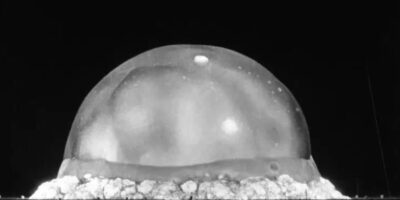 A bola de fogo inicial do teste Trinity do Projeto Manhattan capturada apenas 0.025 segundos (25 milissegundos) após a detonação. Neste momento crítico, a esfera de plasma superaquecido ainda está em contato direto com o solo do deserto, formando um hemisfério perfeito de aproximadamente 100 metros de diâmetro. A temperatura no centro da bola de fogo neste instante atingiu cerca de 10 milhões de graus Celsius, mais quente que a superfície do sol. A luz emitida é tão intensa que iluminou as montanhas circundantes com mais brilho que a luz solar. Esta é a fase de máxima liberação de energia térmica, onde a radiação infravermelha e ultravioleta se propaga em todas as direções à velocidade da luz. Fonte: U.S. Department of Defense.