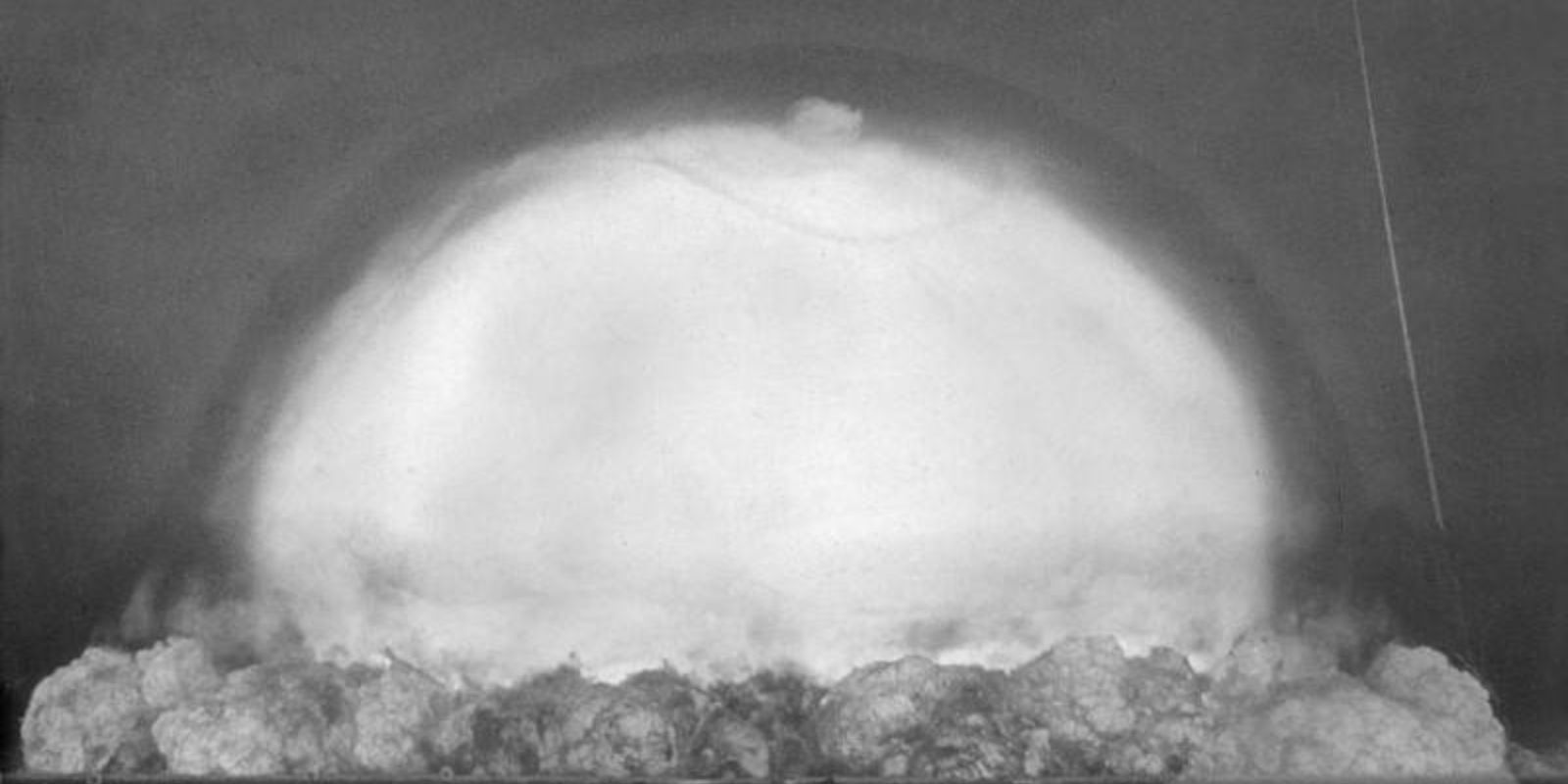 Capturada 0.053 segundos (53 milissegundos) após a detonação, esta imagem mostra a rápida expansão da bola de fogo e o início da formação da onda de choque. A esfera de plasma já se expandiu significativamente, atingindo aproximadamente 200 metros de diâmetro. Observe a base turbulenta onde a bola de fogo ainda interage com o solo do deserto, levantando toneladas de areia e detritos que serão vaporizados instantaneamente. A onda de choque, visível como um anel de compressão atmosférica, se propaga a velocidades supersônicas, comprimindo o ar à sua frente e criando temperaturas extremas. Neste momento, a pressão na frente da onda de choque é de várias atmosferas, suficiente para destruir qualquer estrutura em seu caminho. Fonte: U.S. Department of Defense.