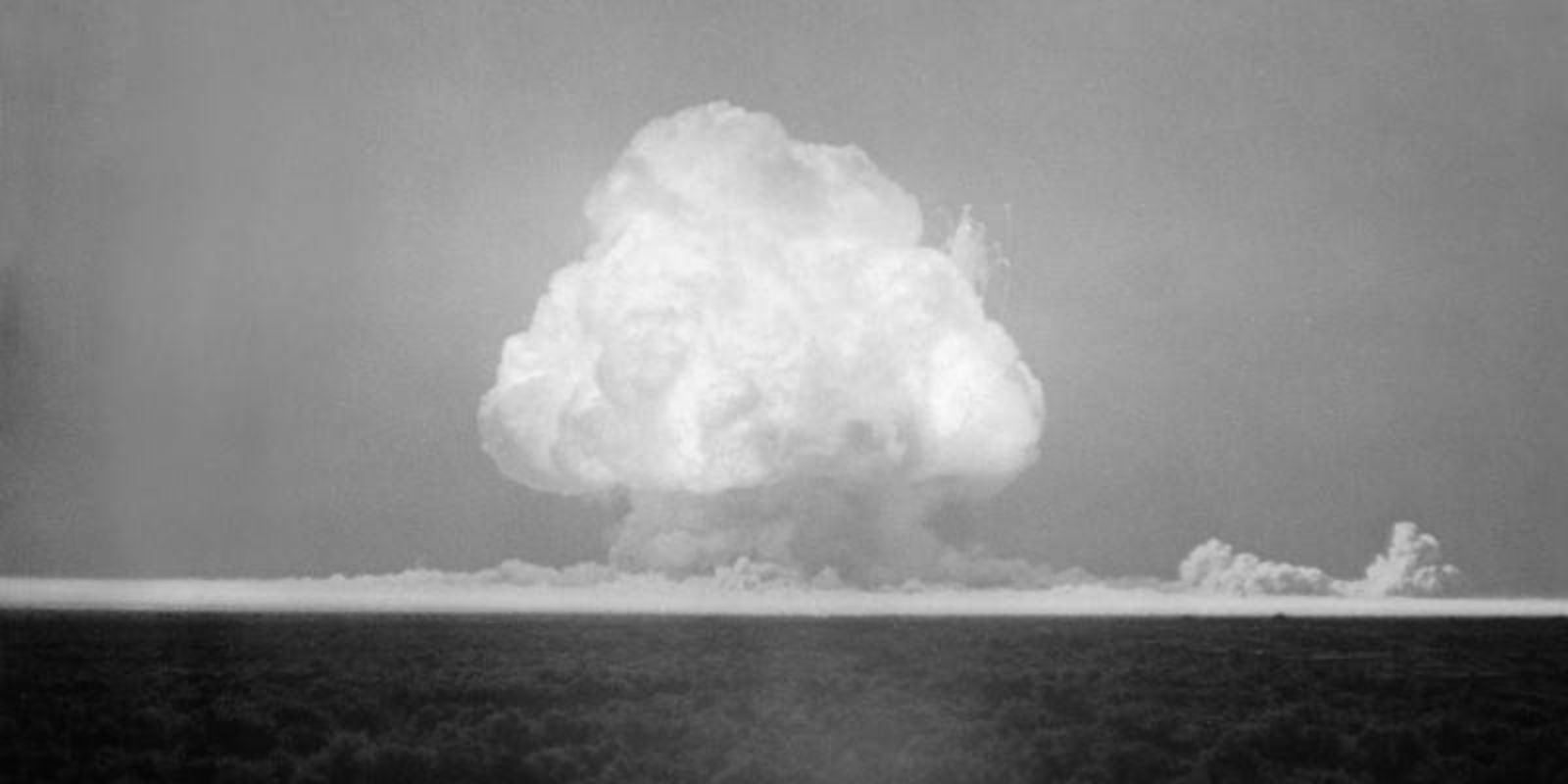 Nove segundos após a detonação, a bola de fogo começa a subir rapidamente devido à convecção térmica, iniciando a formação do icônico cogumelo atômico. A massa de gases superaquecidos, com temperatura ainda na casa dos milhares de graus, ascende através da atmosfera mais fria, criando uma coluna vertical característica. A base da coluna (stem) está se formando, enquanto o topo começa a se expandir horizontalmente ao encontrar camadas de ar mais denso. Neste estágio, a nuvem já atingiu aproximadamente 3 a 4 quilômetros de altura. Os detritos radioativos do solo vaporizado começam a condensar, formando as partículas que darão à nuvem sua aparência característica. A cor esbranquiçada indica a presença de vapor d'água condensado e partículas de poeira radioativa. Fonte: U.S. Department of Defense.