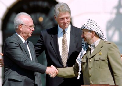 Yitzhak Rabin e Yasser Arafat apertam as mãos no gramado da Casa Branca em 13 de setembro de 1993, observados pelo presidente Bill Clinton. Este momento histórico selou os Acordos de Oslo, que prometiam paz no Oriente Médio através do reconhecimento mútuo entre Israel e a Organização para Libertação da Palestina. O aperto de mão, transmitido ao vivo mundialmente, levou os três líderes a receberem o Prêmio Nobel da Paz em 1994. Fonte: The Philadelphia Inquirer / Vince Musi