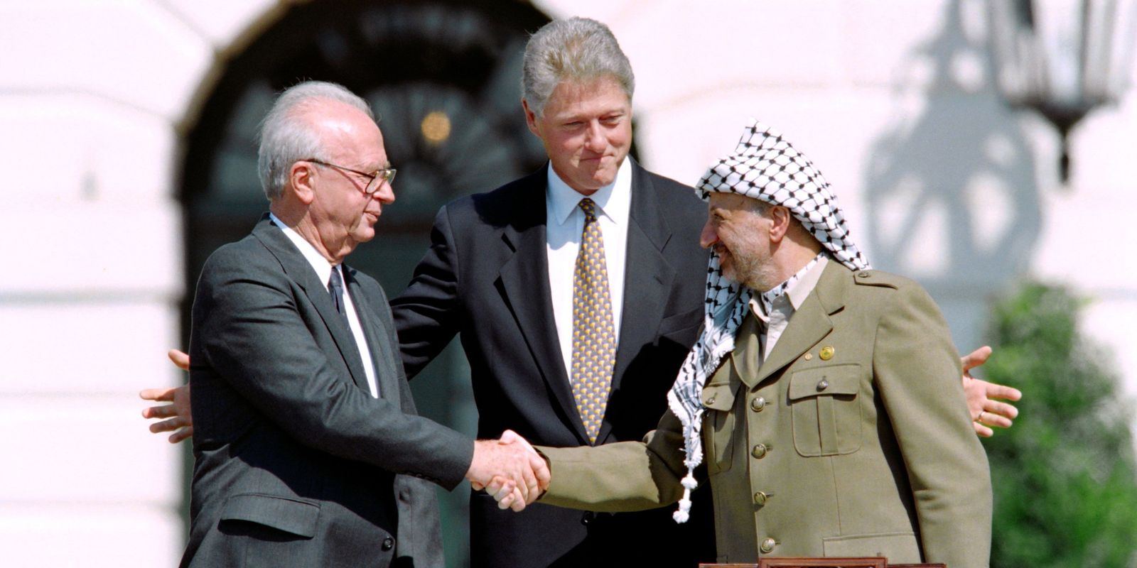 Yitzhak Rabin e Yasser Arafat apertam as mãos no gramado da Casa Branca em 13 de setembro de 1993, observados pelo presidente Bill Clinton. Este momento histórico selou os Acordos de Oslo, que prometiam paz no Oriente Médio através do reconhecimento mútuo entre Israel e a Organização para Libertação da Palestina. O aperto de mão, transmitido ao vivo mundialmente, levou os três líderes a receberem o Prêmio Nobel da Paz em 1994. Fonte: The Philadelphia Inquirer / Vince Musi