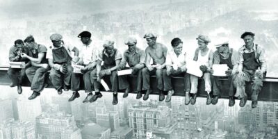 A icônica fotografia "Lunch atop a Skyscraper" de Charles C. Ebbets, tirada em 1932, retrata onze trabalhadores almoçando em uma viga de aço a 256 metros de altura durante a construção do Rockefeller Center. Esta imagem, embora encenada, simboliza a coragem e a resiliência dos trabalhadores da Grande Depressão. Fonte: Rockefeller Center.