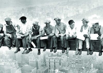 A icônica fotografia "Lunch atop a Skyscraper" de Charles C. Ebbets, tirada em 1932, retrata onze trabalhadores almoçando em uma viga de aço a 256 metros de altura durante a construção do Rockefeller Center. Esta imagem, embora encenada, simboliza a coragem e a resiliência dos trabalhadores da Grande Depressão. Fonte: Rockefeller Center.