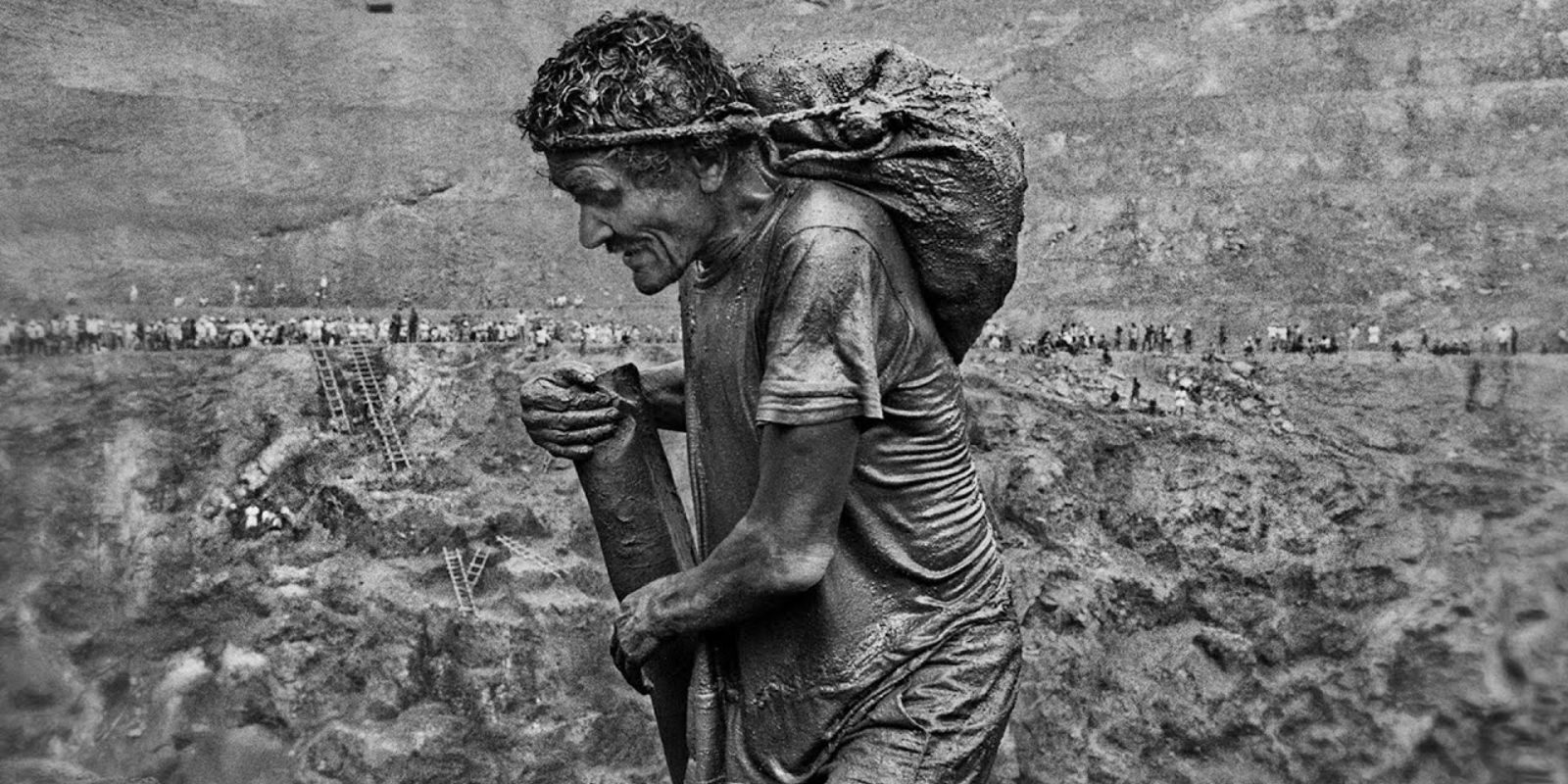 Conhecido como "formiga", um garimpeiro carrega um pesado saco de terra nas costas, subindo as precárias escadas de madeira. A fotografia de Sebastião Salgado é um símbolo do trabalho exaustivo e da esperança incansável de encontrar ouro. Autor: Sebastião Salgado.