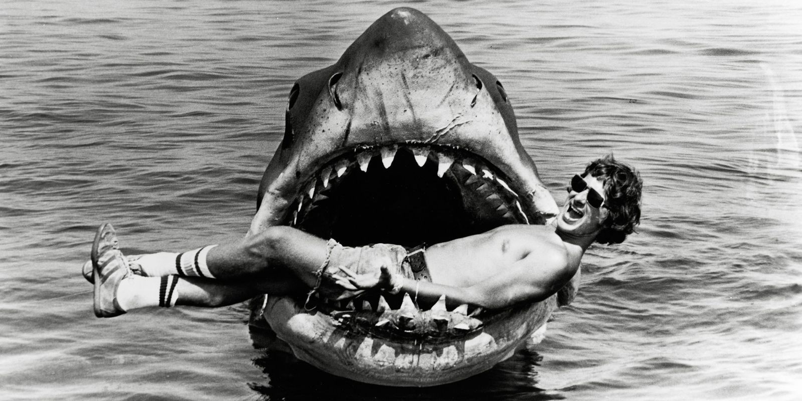 Steven Spielberg posa de forma descontraída na boca do tubarão mecânico Bruce. Esta imagem icônica dos bastidores contrasta com a realidade frustrante das filmagens, onde Bruce afundava regularmente e atrasava a produção por meses. O incidente com George Lucas, que ficou preso nas mandíbulas quando Spielberg travou o mecanismo, mostrou que mesmo em terra o tubarão era imprevisível. Fonte: Universal Pictures / CNN Behind the Scenes