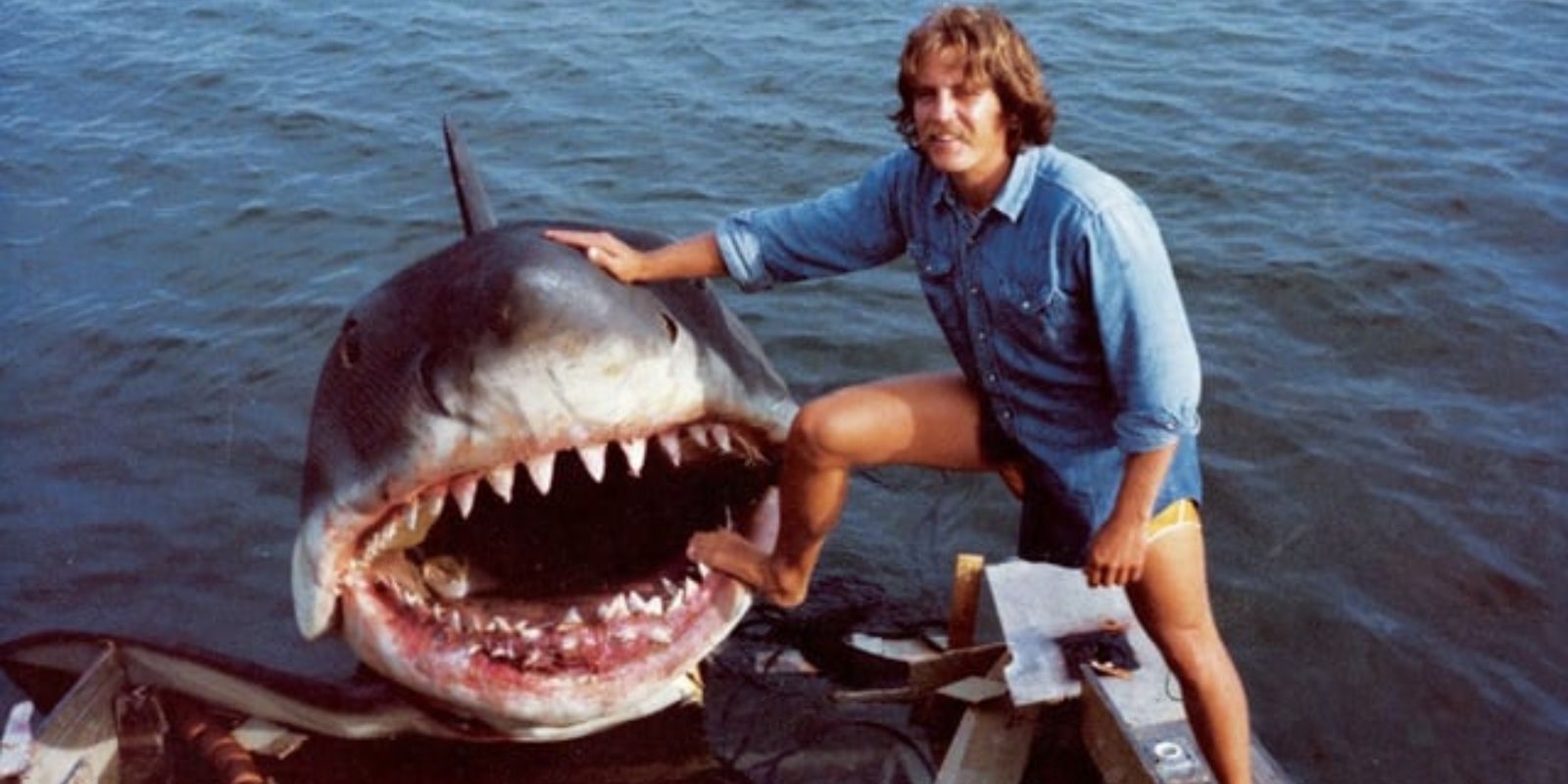 Steven Spielberg sorri durante um momento de descontração nas filmagens de Tubarão em 1974. Apesar da aparência relaxada, o diretor desenvolveu stress pós-traumático que durou anos, dormia no barco de produção e achava que seria demitido todo dia. O fracasso constante de Bruce forçou Spielberg a criar suspense através de câmera subjetiva e da trilha sonora inesquecível de John Williams. Fonte: Universal Pictures / Reddit Moviesinthemaking