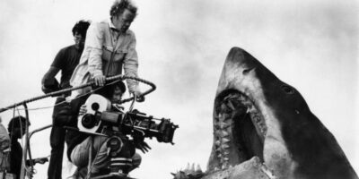 Steven Spielberg dirige uma cena com o tubarão mecânico Bruce durante as filmagens de Tubarão em 1974. O diretor, então com apenas 27 anos, enfrentou desafios monumentais quando Bruce afundava constantemente e seus mecanismos falhavam devido à corrosão da água salgada. Esta fotografia dos bastidores captura um momento raro de funcionamento do tubarão que quase destruiu a produção inteira. Fonte: Universal Pictures / Arquivo de Produção