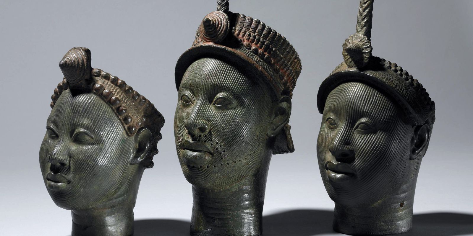 Três cabeças de bronze de Ifé expostas no Museu Britânico, datadas entre 1300-1450. As semelhanças estilísticas entre as esculturas sugerem que foram criadas por um único artista ou em uma única oficina Yoruba. Cada cabeça apresenta características distintas, mas todas compartilham o mesmo nível de sofisticação técnica e realismo naturalista que chocou especialistas europeus quando foram descobertas em 1938. Fonte: © Trustees of the British Museum